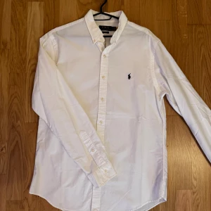 Vit skjorta från Polo Ralph Lauren - Klassisk vit skjorta från Polo Ralph Lauren med broderad logga på bröstet. Skjortan har lång ärm, knappar framtill och krage med knappfäste. Perfekt för dig som gillar stilrena och tidlösa plagg med snygg passform.