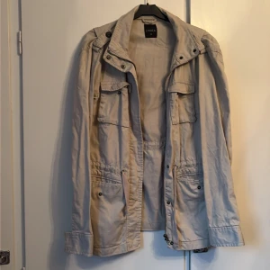 Beige parkajacka från Lindex - Säljer en beige parkajacka från Lindex i storlek 40. Jackan har flera praktiska fickor, snörning i midjan och dragkedja framtill. Materialet är bomull och jackan har en klassisk krage och långärmade ärmar. Perfekt för dig som gillar utility-stil.