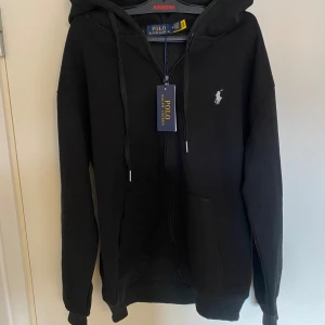 Svart hoodie från Polo Ralph Lauren - Svart hoodie från Polo Ralph Lauren med dragkedja och huva. Klassisk design med vit broderad logga på bröstet och känguruficka framtill. Tillverkad i mjuk bomull och har snörning i huvan. Perfekt för en avslappnad och stilren look.