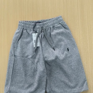 Grå shorts från Polo Ralph Lauren - Snygga grå shorts från Polo Ralph Lauren med elastisk midja och snörning. Klassisk logga broderad på ena benet och mjukt bomullsmaterial som känns skönt mot huden. Perfekta för chill dagar eller när du vill ha en clean och enkel stil.