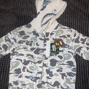 BAPE Shark Camo Hoodie i vitt och grått - Säljer en BAPE Shark Hoodie med ikoniskt hajmotiv på huvan och camouflagemönster i vitt, grått och blått. Hoodien har dragkedja hela vägen, huva och klassisk BAPE-tag. Perfekt för dig som gillar streetwear och vill sticka ut.