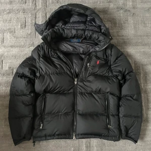 Svart dunjacka Polo Ralph Lauren M - Svart dunjacka från Polo Ralph Lauren med huva, två stora fickor med dragkedja och en mindre bröstficka. Jackan har det klassiska röda Polo-logot på bröstet och är quiltad för extra värme. Perfekt för kalla dagar och har en clean, stilren vibe.