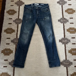 Replay Anbass storlek W30 L32 - Snygga Replay Anbass jeans i slim fit med klassisk femficksdesign. Färgen är mörkblå med slitningar och små hål för en cool, använd look. Jeansen har raka ben och är tillverkade i mjukt denim-material som sitter skönt hela dagen.
