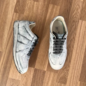 Sköna ljusgrå sneakers med snörning - Snygga sneakers i en unik ljusgrå och vit färg med svart snörning. Klassisk låg modell med rund tå och platt sula. Ovandel i skinn med handmålad finish som ger en cool, vintage look. Perfekta för dig som gillar statement-sneakers med designer-vibe.
