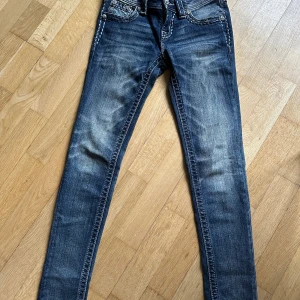 Miss Me skinny jeans med detaljer - Snygga blå Miss Me jeans i skinny fit med coola slitningar och kontrastsömmar. Fickorna har dekorativa knappar och broderade detaljer som ger extra edge. Jeansen har låg midja och klassisk femficksdesign. Perfekt för dig som gillar statement-jeans.