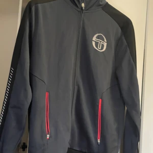 Svart vindjacka från Sergio Tacchini - Svart vindjacka från Sergio Tacchini med röd dragkedja på fickorna och logga på bröstet. Jackan har lång ärm, hög krage och hel dragkedja framtill. Snygga detaljer längs ärmarna och insidan av kragen i rött. Perfekt för sportiga dagar.