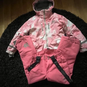 Rosa vinterjacka från 157 Funk - Säljer en rosa vinterjacka från 157 Funk med huva och dragkedja. Jackan har ljusa mönsterdetaljer, flera fickor med dragkedja och justerbara ärmslut. Materialet är syntet och jackan är fodrad för extra värme. Perfekt för kalla dagar och snöaktiviteter.