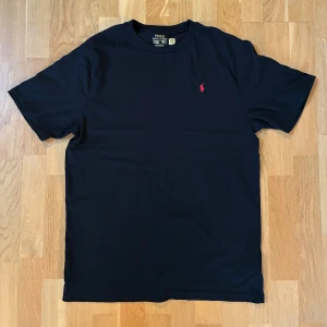 Svart t-shirt från Polo Ralph Lauren - Svart t-shirt från Polo Ralph Lauren med klassisk passform och rund hals. T-shirten har korta ärmar och är tillverkad i mjuk bomull. På bröstet finns den ikoniska röda broderade Polo-loggan som ger en clean och stilren look. Fint skick! Storlek xl(18-20)/170