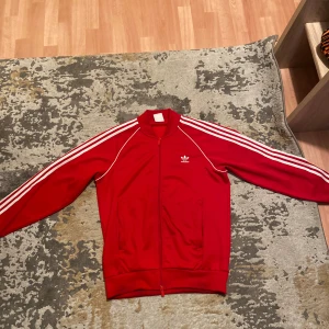 Röd Adidas track jacket med vita ränder - Snygg röd Adidas track jacket med klassiska vita ränder längs ärmarna och Adidas-logga på bröstet. Jackan har hel dragkedja, två fickor fram och ribbade muddar. Perfekt för en sportig och chill look. Storlek L. Säljs för 350kr. Pris kan diskuteras. Endast seriösa köpare.  