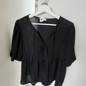 Svart blus med knyt och struktur - Säljer en svart blus med snygg struktur och två knyt framtill. Blusen har korta ärmar och ett lätt, luftigt material som ger en skön känsla. Perfekt för dig som gillar en chill och stilren look med lite extra detaljer.