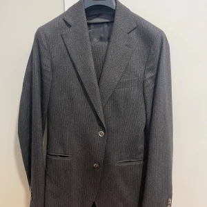 Mörkgrå kritstreck randig  kostym Oscar Jacobson - Snygg mörkgrå kavaj från Oscar Jacobson i exklusiv ull- och kashmirblandning från Tollegno 1900. Kavajen har diskreta ränder, klassisk krage, två knappar framtill och fickor med lock. Perfekt för dig som gillar stilrena och tidlösa plagg. Byxor är storlek 46, kavaj 48. Slim modell