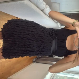 Svart kort klänning med rosett - Säljer en svart kort klänning med coolt texturerad kjol och bred satinmidja med rosett och blommor. Klänningen är ärmlös och har en rundad urringning. Perfekt för dig som vill sticka ut med en unik look.