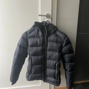 Svart dunjacka Everest - Svart dunjacka från Everest med huva och dragkedja framtill. Jackan är varm och använd fåtal gånger. Säljer då den är för liten 