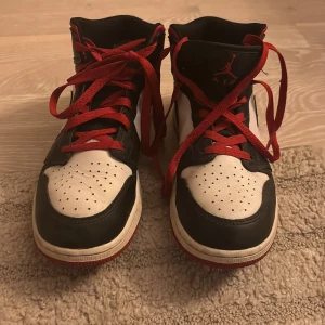 Nike Air Jordan 1 svart/vit/röd - Säljer ett par Nike Air Jordan 1 sneakers i svart, vitt och rött skinn med röda snören och klassisk svart swoosh. Högt skaft och röd yttersula ger en ikonisk look. Perfekta för dig som vill sticka ut med streetstyle och basketvibe. Råka skriva fel på postnummer. 70237