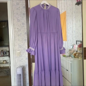 Ljuslila långklänning med pärldetaljer - Superfin ljuslila långklänning med voluminösa långa ärmar och pärlbroderier vid halsringning och ärmslut. Klänningen har en flowy passform, hög hals och vackra volangdetaljer. Perfekt för dig som vill sticka ut med en romantisk och elegant look.