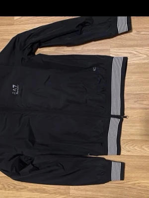 Svart vindjacka från EA7 Emporio Armani - Säljer en svart vindjacka från EA7 Emporio Armani med vita ränder på muddar och nederkant. Jackan har dragkedja framtill och långärmad design. Perfekt för dig som gillar sportig och stilren look.