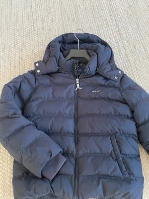 Mörkblå dunjacka från GANT - Säljer en mörkblå dunjacka från GANT med huva och dragkedja framtill. Jackan har broderad logga på bröstet och nacken, samt ribbade muddar vid ärmsluten. Perfekt för kalla dagar och riktigt snygg till jeans eller mjukis. Pris: 600kr. Priset kan diskuteras.