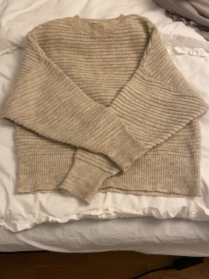 Beige ribbad stickad tröja - Mysig beige stickad tröja med ribbat mönster och rund hals. Tröjan har långa ärmar och breda muddar vid ärmslut och nederkant. 