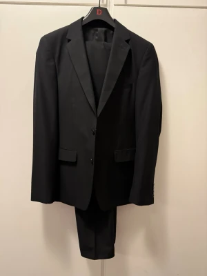 Svart kostym från Dressmann, slim fit - Säljer en svart kostym från Dressmann i slim fit-modell, storlek 50. Kavajen har klassisk krage, två knappar framtill och fickor med lock. Insidan har snygga röda detaljer. Följer även med byxorna på bild storlek 34-32. Kostymen endast använd 2 gånger!! 