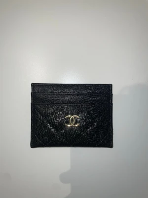 Svart quiltad plånbok från Ch*nel - Säljer en stilren svart plånbok med klassiskt quiltat mönster och guldfärgat CC-logo framtill. Plånboken har flera fack för kort. Perfekt för dig som gillar lyxiga accessoarer med ikonisk design.