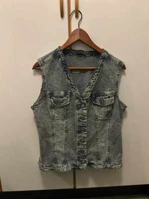 Grå jeansväst med knappar - Säljer en snygg grå jeansväst med slitna detaljer och v-ringning. Västen har två bröstfickor med lock och metallknappar framtill. Perfekt att slänga över en t-shirt för en cool look.