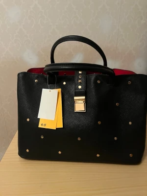 Svart handväska med gulddetaljer - Snygg svart handväska med guldfärgade nitar och spänne framtill. Väskan har två handtag, dragkedja och röd insida som ger en cool kontrast. Tillverkad i skinnimitation med struktur och har plats för allt du behöver till vardagen.