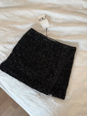 Svart paljettkjol från Zara - Säljer en svart kortkjol från Zara täckt med glittriga paljetter. Kjolen har en rak passform och en liten slits 