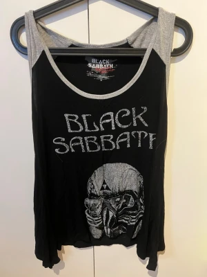 Black Sabbath linne med dödskalle - Säljer ett svart linne från Black Sabbath med gråa kanter och tryck framtill. Framsidan har ett stort dödskallemotiv och texten 'Black Sabbath' i vintage-stil. Baksidan har en liten logga upptill. Perfekt för dig som gillar rock och bandmerch. Oversized 