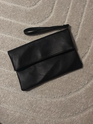 Svart kuvertväska i skinnimitation - Stilren svart kuvertväska med handledsrem i skinnimitation. Enkel och klassisk design utan synliga detaljer eller mönster. Perfekt storlek för det viktigaste och lätt att bära med sig.