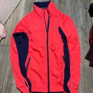 Rosa vindjacka från SOC, strl 34 - Säljer en sportig rosa vindjacka från SOC i storlek 34. Jackan har mörkblå detaljer längs ärmar och sidor, hel dragkedja framtill och två fickor med dragkedja. 