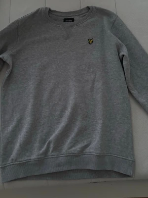 Grå sweatshirt från Lyle & Scott - Klassisk grå sweatshirt från Lyle & Scott med rund halsringning och den ikoniska gula loggan broderad på bröstet. Tröjan har ribbade muddar vid ärmslut och nederkant samt är långärmad. Perfekt för en chill och stilren look.