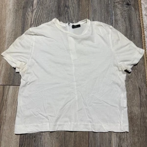 Vit t-shirt/croptop med dragkedja från Zara - Enkel och stilren vit t-shirt/crop top  från Zara med rak passform och rund hals. T-shirten har korta ärmar och en diskret dragkedja i nacken som ger en cool detalj. Perfekt till jeans eller kjol för en clean look. ‼️ obs att det är strl L på lappen men jag skulle säga S 