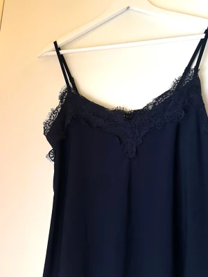 Mörkblå linnetopp med spets -  Vackert marinblått linne med spets vid ringningen ovanför bysten. En feminin och tidlös topp som funkar lika bra till vardags som till mer uppklädda tillfällen.