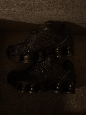 Svarta Nike Shox sneakers - Säljer ett par svarta Nike Shox sneakers med ikoniska fjädrande sulor och meshdetaljer. Skorna har snörning, diskreta röda loggor på plösen och coola orange detaljer under sulan. Perfekta för dig som vill ha en sportig och futuristisk look.