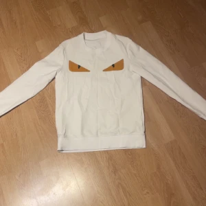 Vit Fendi tröja - Säljer en vit Fendi tröja med långa ärmar och ett par coola ögon i gult och svart tryckta på bröstet. Tröjan har rund halsringning och är gjord i mjukt bomullsmaterial. Perfekt för dig som vill sticka ut lite extra.