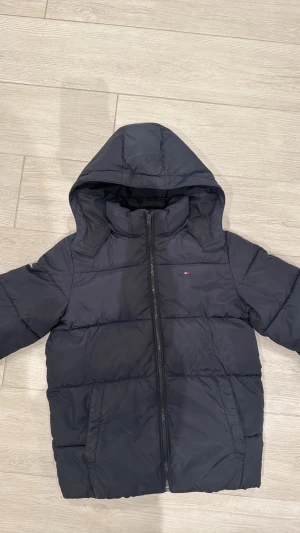 Svart pufferjacka från Tommy Hilfiger - Säljer en svart pufferjacka från Tommy Hilfiger med huva och dragkedja. Jackan har broderad logga på bröstet och ärmen samt stor logga på insidan av kragen. Tillverkad i syntet sopass att den är riktigt varm och perfekt för kalla dagar. Storleken är 16-18 år fast jag skulle säga att den e ganska liten i storlek och passar runt 14-15 år