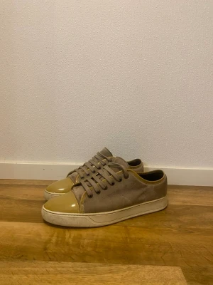 Beige sneakers från Lanvin i mocka - Säljer ett par beige sneakers från Lanvin med ovandel i mocka och tåparti i glansigt skinn. Skorna har rund tå, snörning och vit platt sula. Snygg kontrast mellan material och färg, perfekta till jeans eller chinos.