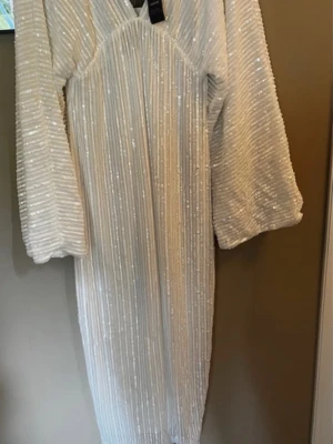 Vit paljett långklänning med v-ringning -  vit långklänning med glittriga paljetter och v-ringad hals. Klänningen har långa ärmar och ett randigt mönster av paljetter. Oanvänd 