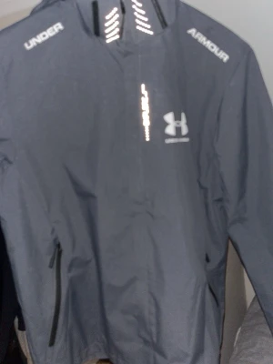 Grå vindjacka från Under Armour - Snygg grå vindjacka från Under Armour med logga på bröstet och text på axlarna. Jackan har dragkedja framtill och vid fickorna, samt reflekterande detaljer på kragen. Perfekt för vindiga dagar.