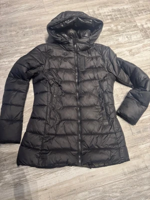 Svart jacka från G-Star Women - Svart jacka från G-Star Women i storlek L. Jackan har huva, dragkedja framtill och två fickor med knappar. Den är quiltad och har en längre passform som ger extra värme. Perfekt för kalla dagar och har en stilren look.