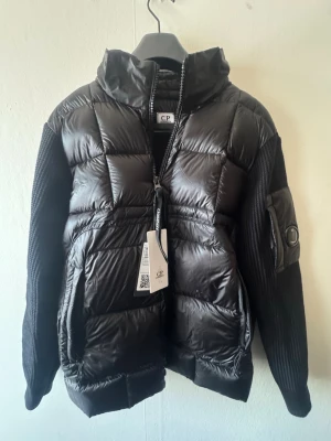 Svart pufferjacka från C.P. Company - Säljer en svart pufferjacka från C.P. Company med stickade ärmar och vadderad framsida. Jackan har dragkedja framtill, hög krage och en ikonisk detaljficka med glaslins på ena ärmen. Perfekt för dig som vill ha en modern och sportig look.