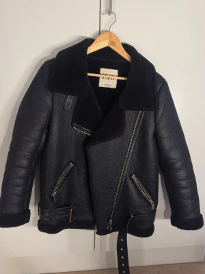 Vinterjacka från Zara  - Säljer en svart teddyjacka från Zara med bred krage, dragkedjor och bälte i midjan. Jackan har en cool bikerstil med grov struktur, Perfekt för kalla dagar 