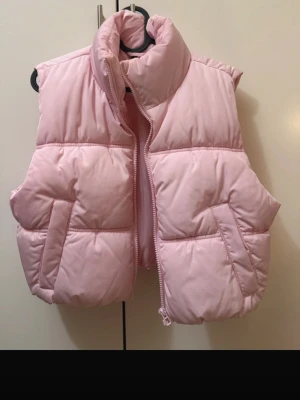 Ljusrosa dunväst med hög krage - Säljer en ljusrosa dunväst med hög krage och dragkedja framtill. Västen har två snedställda fickor och quiltad design som ger en puffig look. Perfekt att slänga över en hoodie eller tröja för extra värme. Enkel och stilren modell.