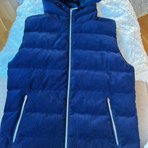 Blå dunväst från Zadolo - Snygg blå dunväst från Zadolo med huva och två fickor med dragkedja. Västen är quiltad och har ett yttertyg i manchester som ger en cool och modern look. Perfekt att slänga över en hoodie när det är kyligt ute.