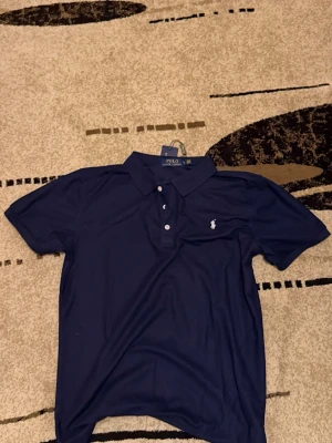 Marinblå pikétröja Polo Ralph Lauren - Säljer en klassisk marinblå pikétröja från Polo Ralph Lauren. Tröjan har krage, knäppning med tre vita knappar och den ikoniska broderade loggan på bröstet. Tillverkad i mjuk bomull som känns skön mot huden. Perfekt till jeans eller shorts.
