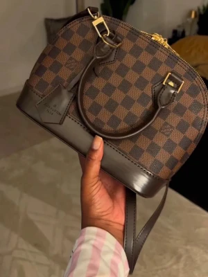 handväska  - Säljer en ikonisk  handväska i brunt Damier-mönster. Väskan har rundad form, två handtag, avtagbar axelrem och guldfärgade metalldetaljer. Utsidan är i canvas och skinn, insidan är röd textil. Perfekt för dig som vill ha en klassisk designerbag.