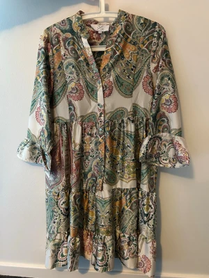 Mönstrad långklänning från JS - Säljer en bohemisk långklänning från JS med färgstarkt paisleymönster i grönt, gult, rosa och beige. Klänningen har v-ringning, knappar framtill och volangdetaljer vid ärmslut. Materialet känns lätt och luftigt, perfekt för varmare dagar.