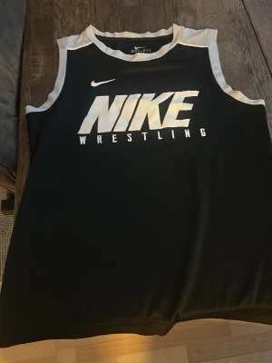 Nike Dri-Fit Wrestling Linne Svart S - Snyggt svart linne från Nike med stora vita loggan och texten 'Wrestling' framtill. Tillverkat i Dri-Fit material som andas och håller dig torr. Kontrastfärgade vita kanter vid ärmhål och halsringning. Perfekt för träning eller brottning.