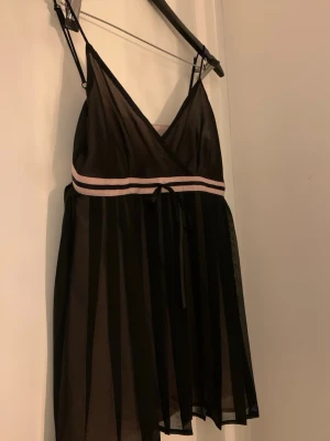 Svart kort omlottlinne - Säljer en svart linne med omlottdesign och tunna axelband.  Linnet har ett rosa band i midjan med en liten rosett framtill! Vintage Oasis! 