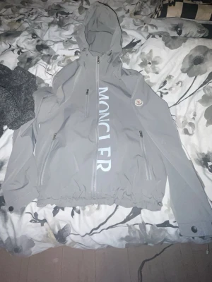 Grå vindjacka från Moncler - Säljer en grå vindjacka från Moncler med stor logga längs dragkedjan och klassisk patch på ärmen. Jackan har dragkedja framtill och elastiska muddar. Perfekt för dig som vill ha en clean och stilren look. Plagget är s men passar delvis m.